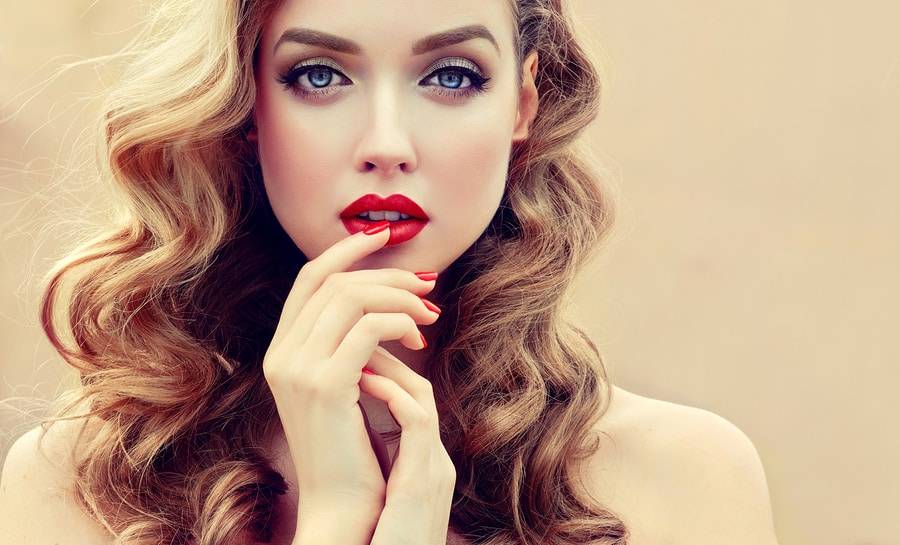 Beautiful blonde model girl with long curly hair . Hairstyle wavy curls . Red lips and nails manicure . Fashion , beauty and make up portrait