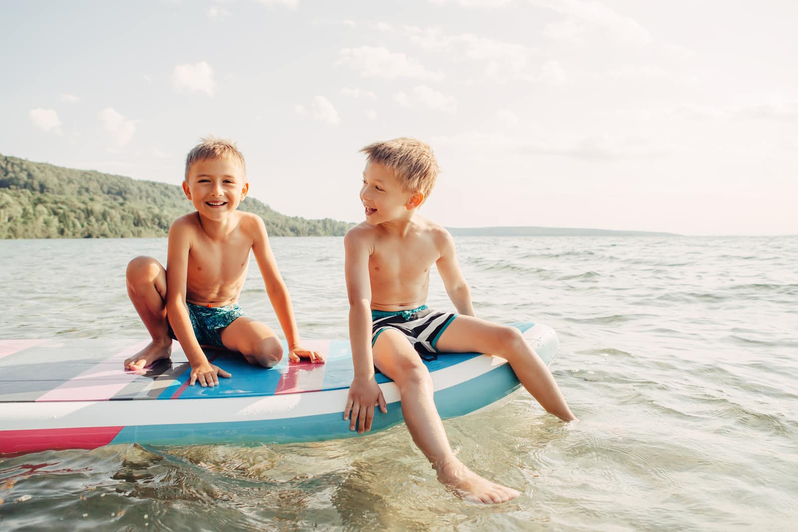 Two smiling Caucasian boys kids sitting on paddle sup surfboard in water