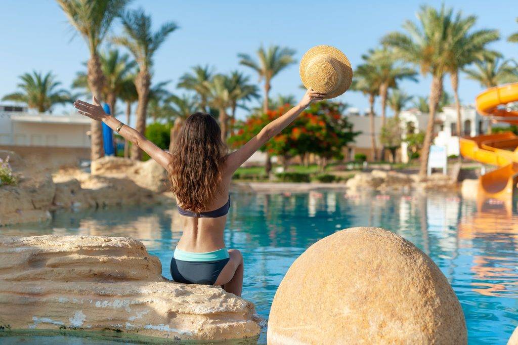 Woman sits on stone near of the swimming pool and rise her hands holding hat. Beautiful exotic hotel relax with palms in Egypt. Blue water texture is place for your text